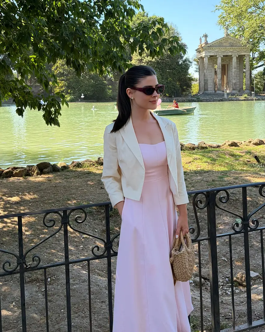 Stephanie de vestido rosa e casaco claro, à beira de um lago com um pequeno templo ao fundo